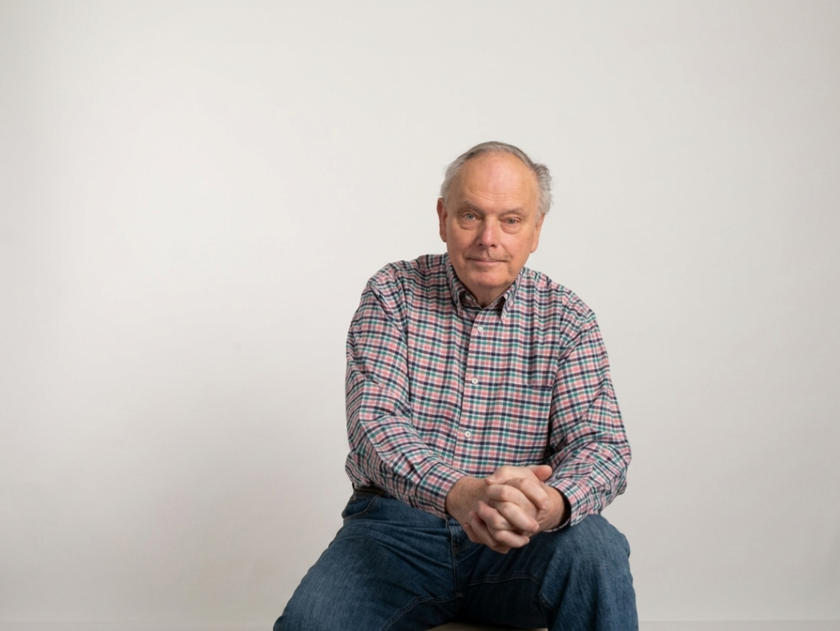 A person sits facing the camera against a light background, wearing a plaid button-down shirt with hands clasped.