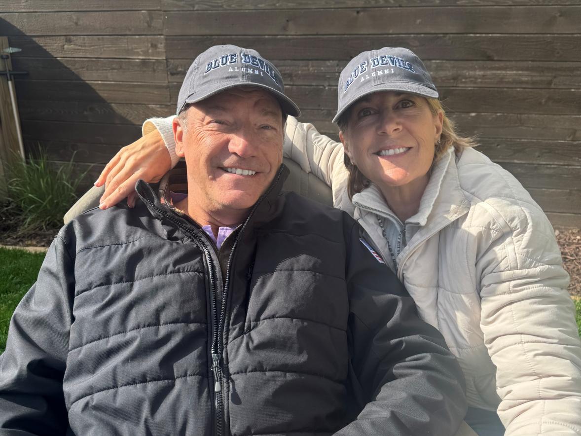 Two adults wearing “Blue Devils Alumni” hats sit outdoors on a bench, leaning together for a close-up photo with a wooden fence and greenery behind them.