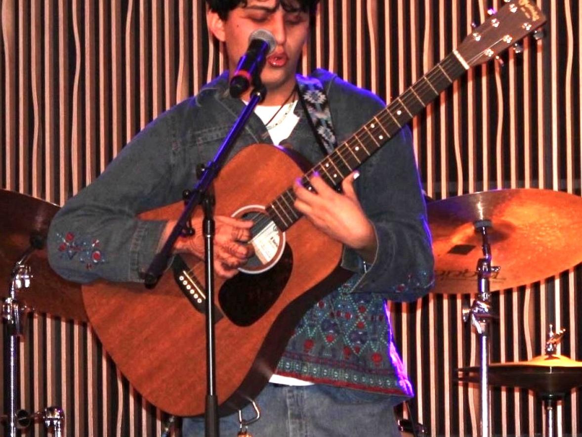 A student performs on stage, singing into a microphone while playing an acoustic guitar, with a drum set visible in the background.