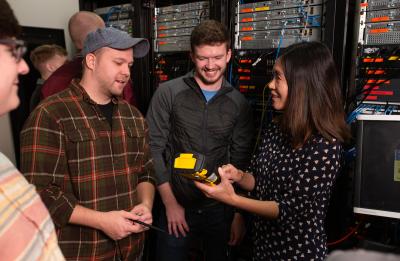 Students in Holly Yuan's capstone course pose with various networking equipment in a lab in Micheels Hall.