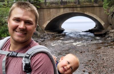 Chris Ester stands in front of a bridge, smiling at the camera. A child in a backpack leans over his shoulder and smiles as well.