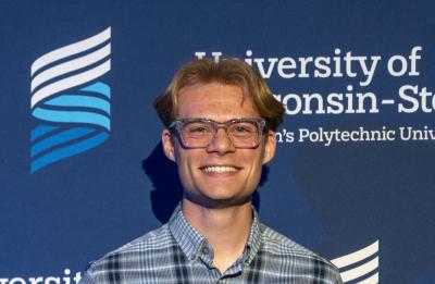 A person stands in front of an official University of Wisconsin–Stout backdrop while holding a clear award plaque. They are wearing a plaid shirt and slacks.