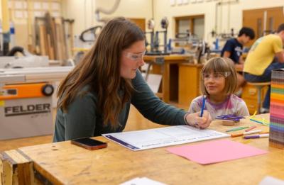 An adult helps a young child with a drawing or writing activity at a workbench in a woodshop classroom. Colored pencils, paper and tools are scattered on the table, and large woodworking equipment is visible in the background.