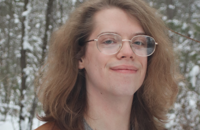 Outdoor winter portrait A person with long, wavy hair stands outside in a snowy forest, wearing glasses, a brown leather jacket, and a gray henley shirt.