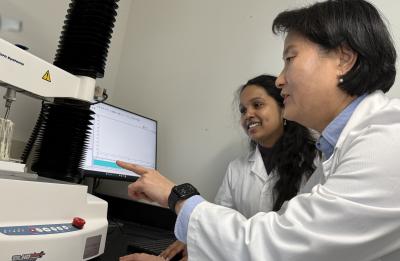 Two people in lab coats work together at a laboratory texture analyzer. One gestures toward a computer screen displaying data while the other stands nearby. The machine and surrounding lab equipment are clearly visible.