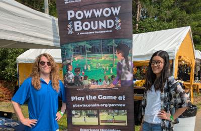 Two people stand on either side of a tall “Powwow Bound” promotional banner at an outdoor event with vendor tents and trees in the background.