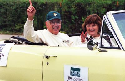Two adults riding in a light-colored convertible during a parade; one waves while a sign on the car door reads, “Jim Bensen, President, Bemidji State University.”