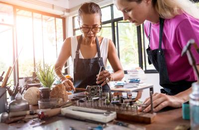 One woman with glasses and an apron is using a hammer and punch on a metal dapping block, while the other woman in a pink shirt and apron watches closely. The workbench is cluttered with various specialized tools, including a tray of rotary burrs and polishing bits, in a bright workshop with large windows in the background.