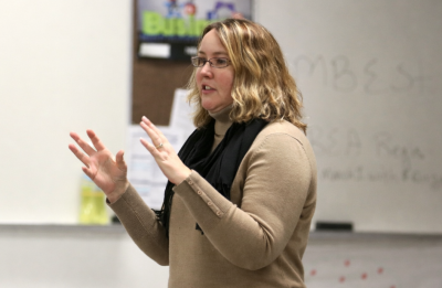 A person stands at the front of a classroom with hands raised mid-gesture. The individual is wearing a light-colored long-sleeve top and a dark scarf. Behind them is a whiteboard with faint writing and a bulletin board displaying classroom materials.
