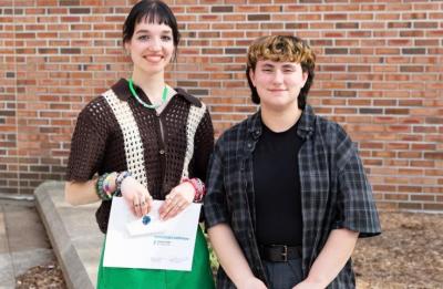 Two people stand side by side outdoors in front of a brick wall. One holds an envelope labeled “Stout University Foundation Scholarship.”