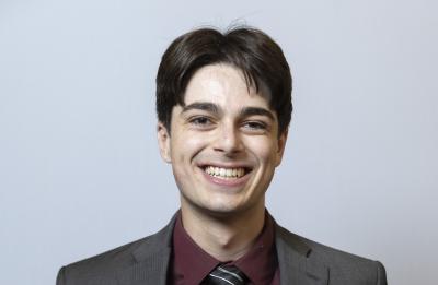 A person stands against a light gray background wearing a dark gray suit, maroon dress shirt and striped tie, facing the camera with hands in pockets.