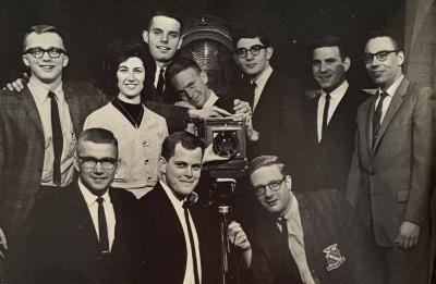 Black-and-white group photograph of eleven students standing and kneeling around a large, box-style camera on a tripod. The students are dressed in suits, ties and cardigans, typical of the mid-20th century, and pose closely together in an indoor studio or classroom setting. A large circular light or reflector is visible behind them.