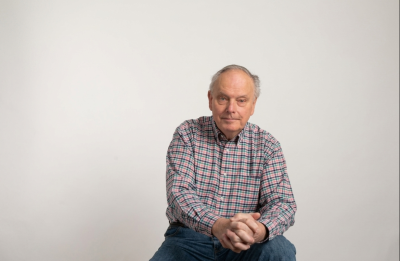 A person sits facing the camera against a light background, wearing a plaid button-down shirt with hands clasped.