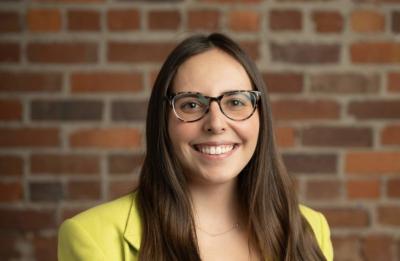 Professional headshot of a person with long dark hair wearing glasses, a bright green blazer, and a white top, standing in front of a brick wall and facing the camera.