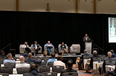 A conference panel takes place on a stage with four seated speakers and one presenter at a podium, facing an audience seated at round tables in a large ballroom.