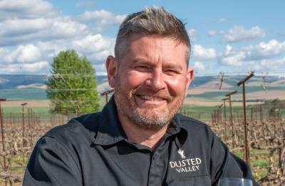Person standing in a vineyard holding a glass of white wine, with rows of vines and rolling hills in the background.