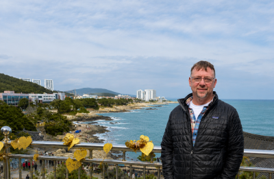 A person stands along a coastal overlook with ocean waves, rocky shoreline and buildings visible in the background.