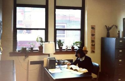 A person sits at a desk reading a book in an office with large windows, plants and a desk lamp.