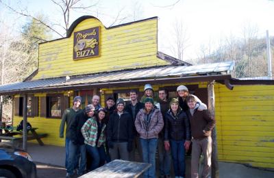 Group of people standing outside a weathered yellow building with a sign reading “Miguels Pizza,” with trees and a picnic table nearby.