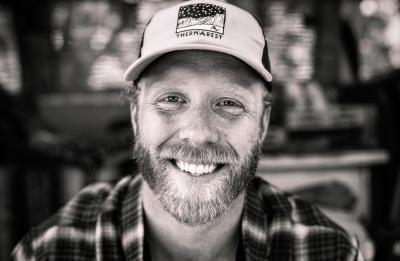 Close-up black-and-white portrait of a person with a beard wearing a cap, smiling toward the camera.