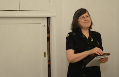A person speaks at the front of a room, holding notes and standing beside a door in a simple indoor setting.