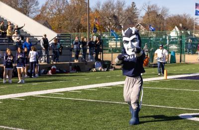 UW-Stout Polytechnic mascot, Blaze, the Blue Devil, stands on the football field pointing forward.