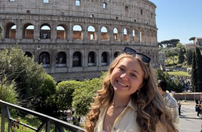 Person smiling in front of the Colosseum in Rome on a sunny day with visitors and greenery in the background.