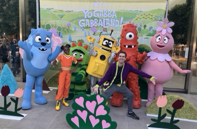 Michael Hilliger poses with costumed characters from Yo Gabba GabbaLand! in front of a promotional display featuring flowers and a colorful backdrop.