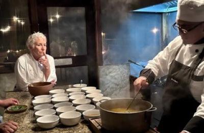 Chef stirring a large pot while preparing food in a kitchen as another person watches nearby.
