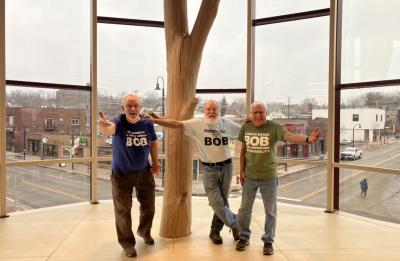 Three visitors wearing matching “BOB” T-shirts pose beside a large wooden tree sculpture inside a museum, with floor-to-ceiling windows overlooking a downtown street.