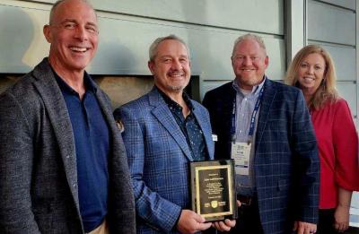 Four people stand together indoors while one person holds a recognition plaque during an award presentation.