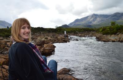 Julia sits on rocks beside a flowing river with mist-covered mountains and cloudy skies in the background.