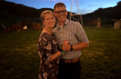 Two people pose together outdoors at dusk beneath string lights.