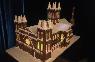 A detailed gingerbread replica of a large cathedral-style building sits on display against a dark backdrop. The structure features multiple towers, snow-covered rooftops and icicle-like frosting along the edges. Illuminated stained-glass-style windows glow in yellow, red and green, adding warmth and intricate detail to the festive architectural design.