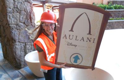 A person wearing a hard hat, safety glasses and a reflective vest holds an Aulani Resort sign during a construction or facilities project.