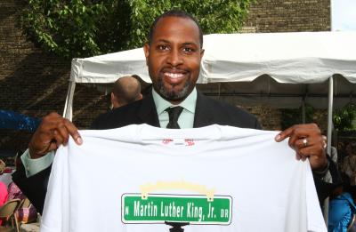 Person in a dark suit and tie holds up a white T-shirt printed with a green street sign reading “N Martin Luther King, Jr. Dr” at an outdoor event under a white tent.