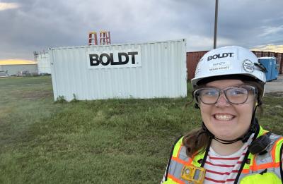 Megan wears construction safety gear and a hard hat at a job site, standing near a BOLDT project trailer under cloudy skies during an internship experience.