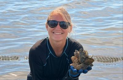 Person in chest waders and gloves kneels on a shoreline beside calm water, holding several oysters while surrounded by oyster shells and restoration equipment.