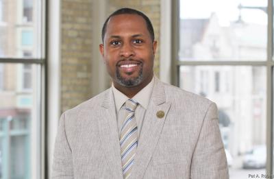 Professional headshot of a person wearing a light-colored suit jacket, striped tie and collared shirt, standing indoors in front of large windows with an urban streetscape visible in the background.