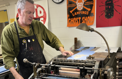 An artist adjusts paper on a vintage letterpress printing press in a studio displaying colorful poster prints on the wall.