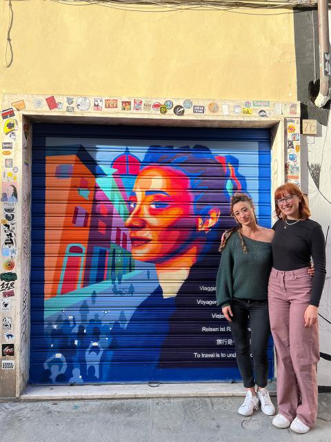 Two people stand in front of a mural of the portrait of a women