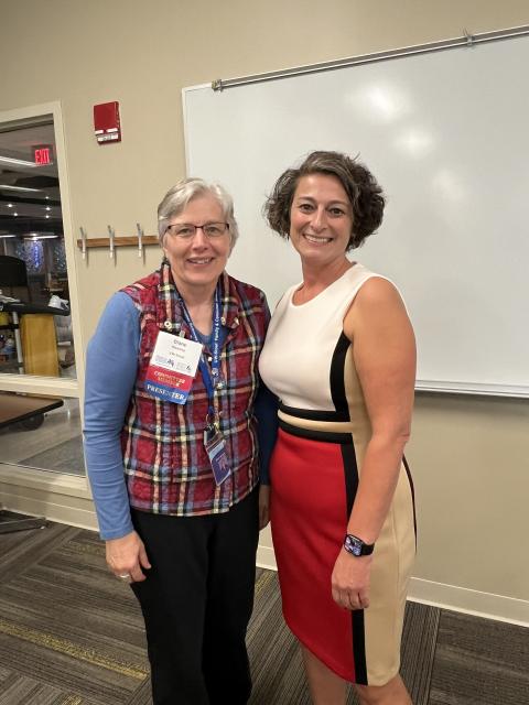 Two people stand side by side in a classroom or meeting room. One person is wearing a blue long-sleeve shirt with a red and blue plaid vest and conference name badges. The other person is wearing a sleeveless dress with beige, black, and red color blocking and a smartwatch. They are standing in front of a whiteboard, with a coat rack and a doorway visible in the background.