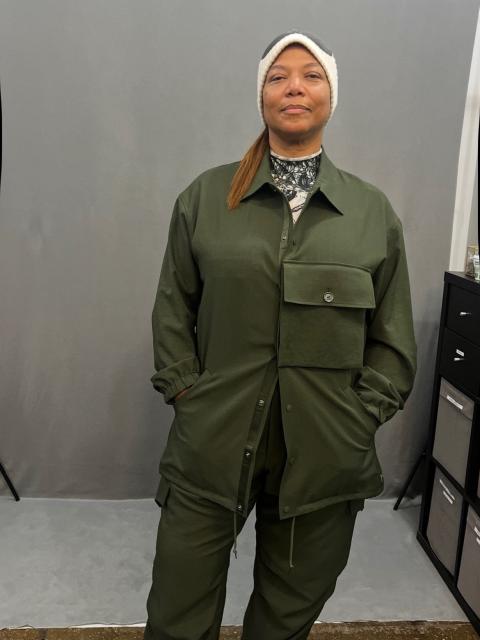 Person wearing a dark green utility-style outfit with a button-up jacket featuring a large flap pocket and matching cargo-style pants. Under the jacket is a black-and-white patterned top. The individual has a white headband and black shoes on one foot and a multicolored sneaker on the other, standing on a brown textured floor against a gray backdrop.