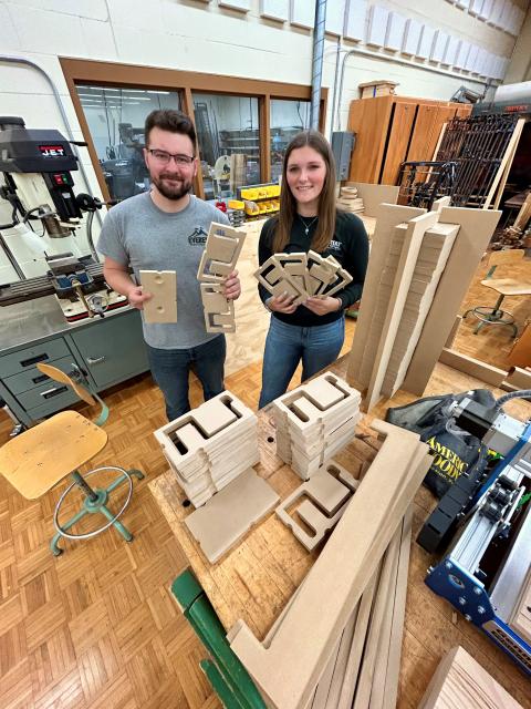 Two students stand in a woodshop holding multiple cut wood pieces shaped for a project. Stacks of similar wood components are arranged on the workbench in front of them, with woodworking tools and machinery surrounding the space.