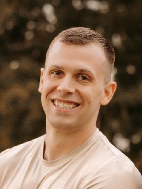 A person stands outdoors wearing a light-colored T-shirt. Trees and greenery form a soft, blurred background behind them.