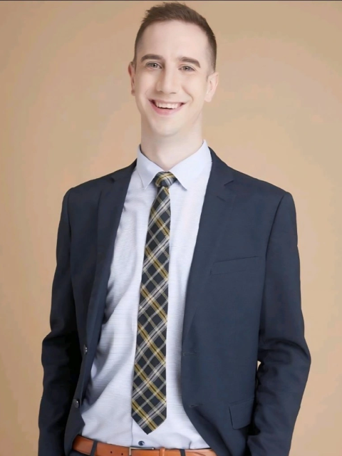 A person wearing a dark blazer, patterned tie, light-colored dress shirt, and brown belt stands against a plain beige background. The individual is posed with hands in pockets, and the outfit is neatly arranged and professional in appearance.