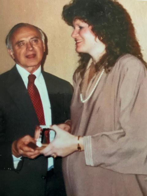 Two people standing indoors, dressed in formal clothing, with one person handing an award or boxed item to the other. The background consists of a plain wall with warm lighting, suggesting an event or ceremony setting.