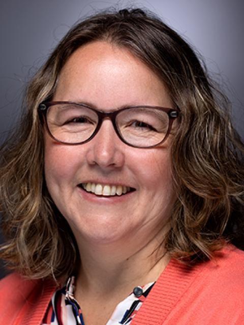 A close-up portrait of a person with shoulder-length wavy hair, wearing a light-colored cardigan over a patterned shirt. A neutral studio backdrop is behind them.