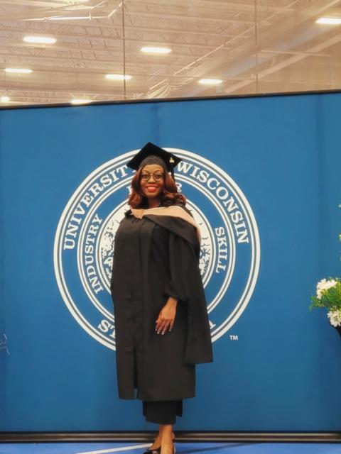 Graduate wearing a black cap and gown stands in front of a University of Wisconsin–Stout backdrop inside a fieldhouse,