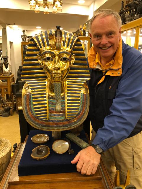 A person stands beside a gold Egyptian pharaoh bust displayed on a pedestal inside a shop filled with artifacts and sculptures.